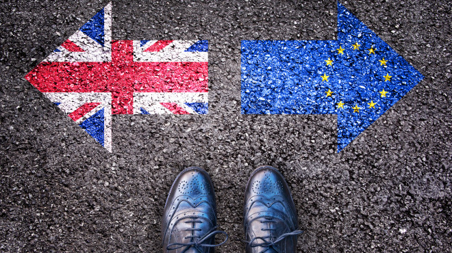 Zwei Füsse stehen vor zwei Pfeilen. Nach links zeigt der Pfeil mit Grossbritanniens Flagge, nach rechts der Pfeil mit der EU Flagge