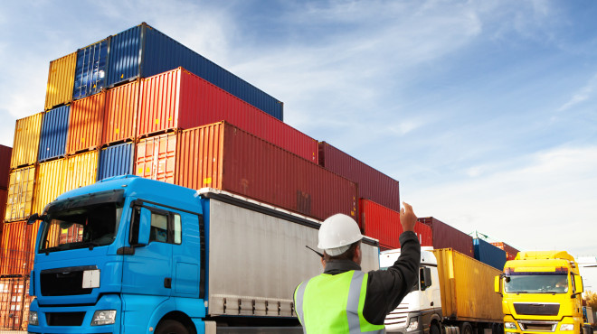 Arbeiter lotst Lastwagen, im Hintergrund Frachtkontainer