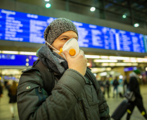 Mann mit Schutzmaske steht am Bahnhof vor einer digitalen Anzeigetafel
