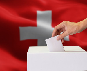 Hand wirft Wahlzettel in Wahlbox ein, im Hintergrund eine Schweizerflagge